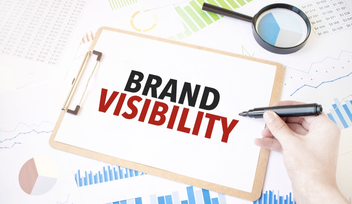 Hand writing "Brand Visibility" on a clipboard surrounded by analytics charts, financial reports, and a magnifying glass — measuring how brands show up in AI search
